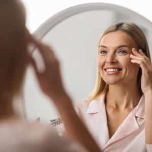 Cheerful mature woman looking at mirror at home, applying eye cream. Attractive middle aged lady using nourishing serum for eye zone while sitting at dressing table in bedroom, selective focus