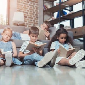 three children are engrossed in reading books on a stylish, sunlit floor in a cozy, modern living room. A relaxed adult reclines on a sofa nearby. The warm tones and contemporary decor create a welcoming and peaceful atmosphere, perfect for family bonding and quiet moments.