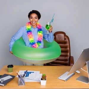 Young African American woman in formal shirt wearing a lei and holding a drink, with a buoy at office desk, on grey background.