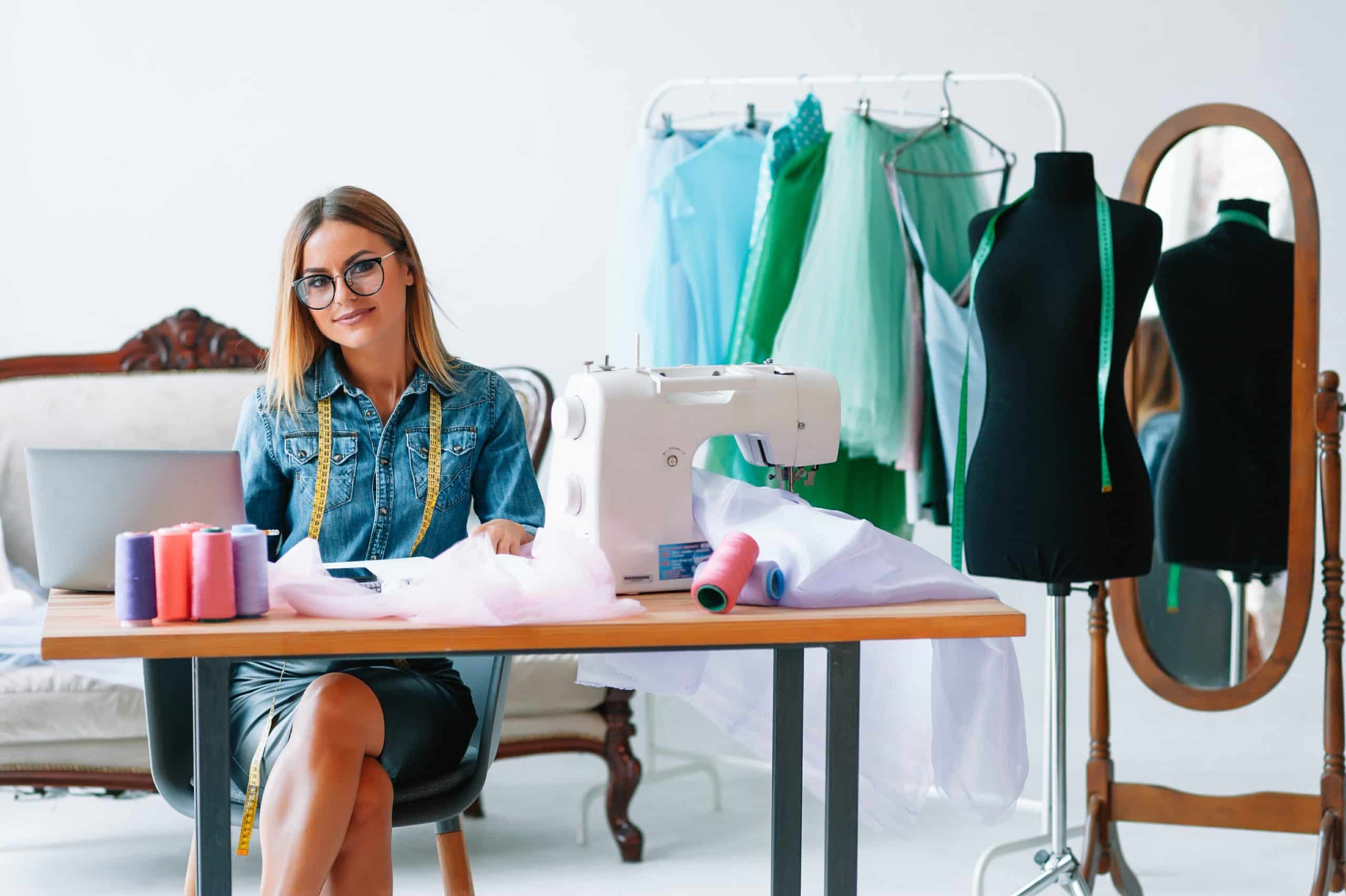 Smiling fashion designer looking at camera at workplace, dressmaker, needlewoman or tailor shop owner sitting at desk with color swatches pantone and embroidery design sketches on the wall, portrait