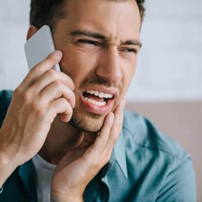 Young man talking by smartphone and looking away while suffering from tooth pain at home