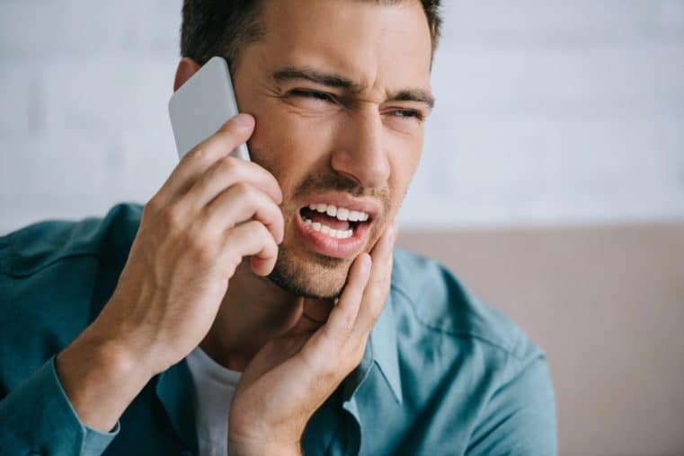 Young man talking by smartphone and looking away while suffering from tooth pain at home