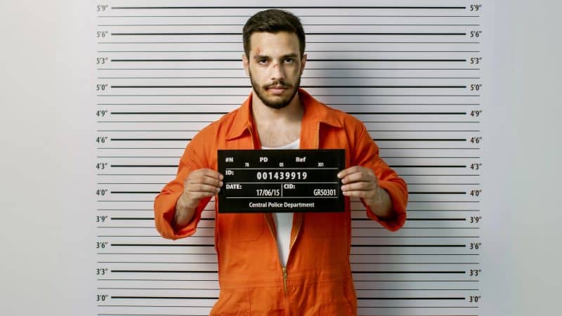 In a Police Station Arrested Man Getting Front-View Mug Shot. He's Wearing Prisoner Orange Jumpsuit and Holds Placard. Height Chart in the Background.