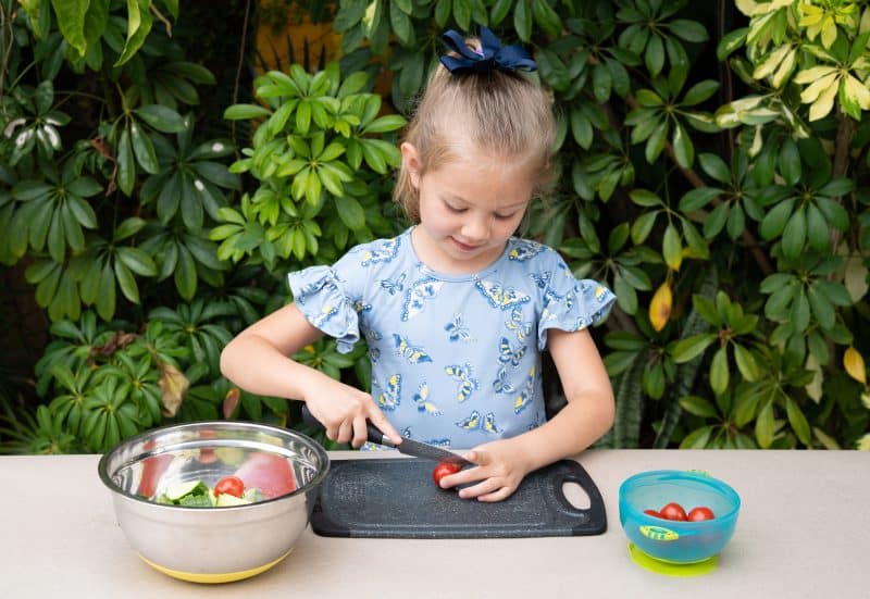 A cheerful young girl is learning to slice a fresh tomatoes on a black cutting board outdoors. Surrounded by lush greenery, she engages in a hands-on cooking activity, promoting healthy eating habits Cheerful Young Girl Learning Slice Fresh Tomatoes Black Cutting Board Outdoors Surrounded Lush Greenery She Engages Hands Cooking Activity Promoting Healthy Eating Habits